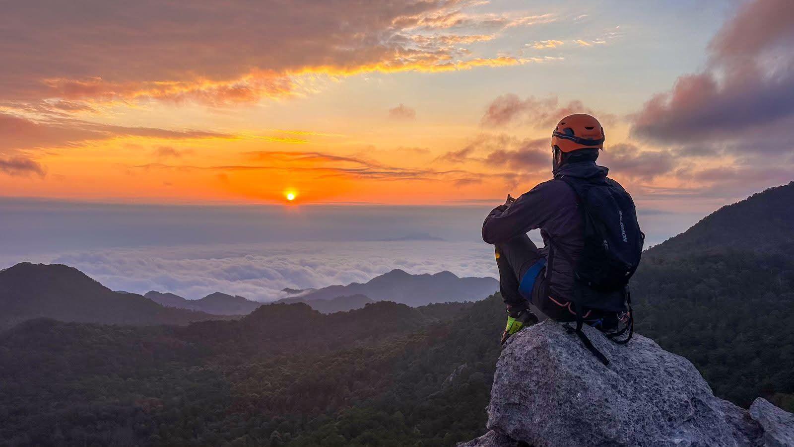 Excursionista sentado en una roca de la cima, de espaldas, frente al amanecer sobre las nubes