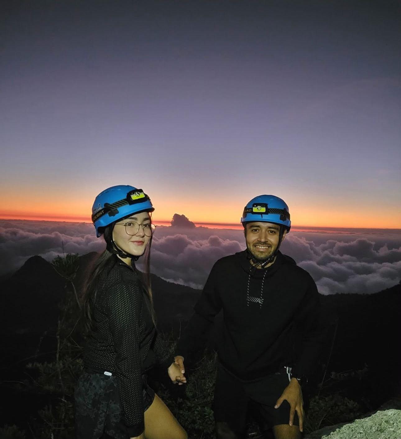 Pareja con casco y lámpara frontal en la cima, con el mar de nubes y el cielo morado detrás