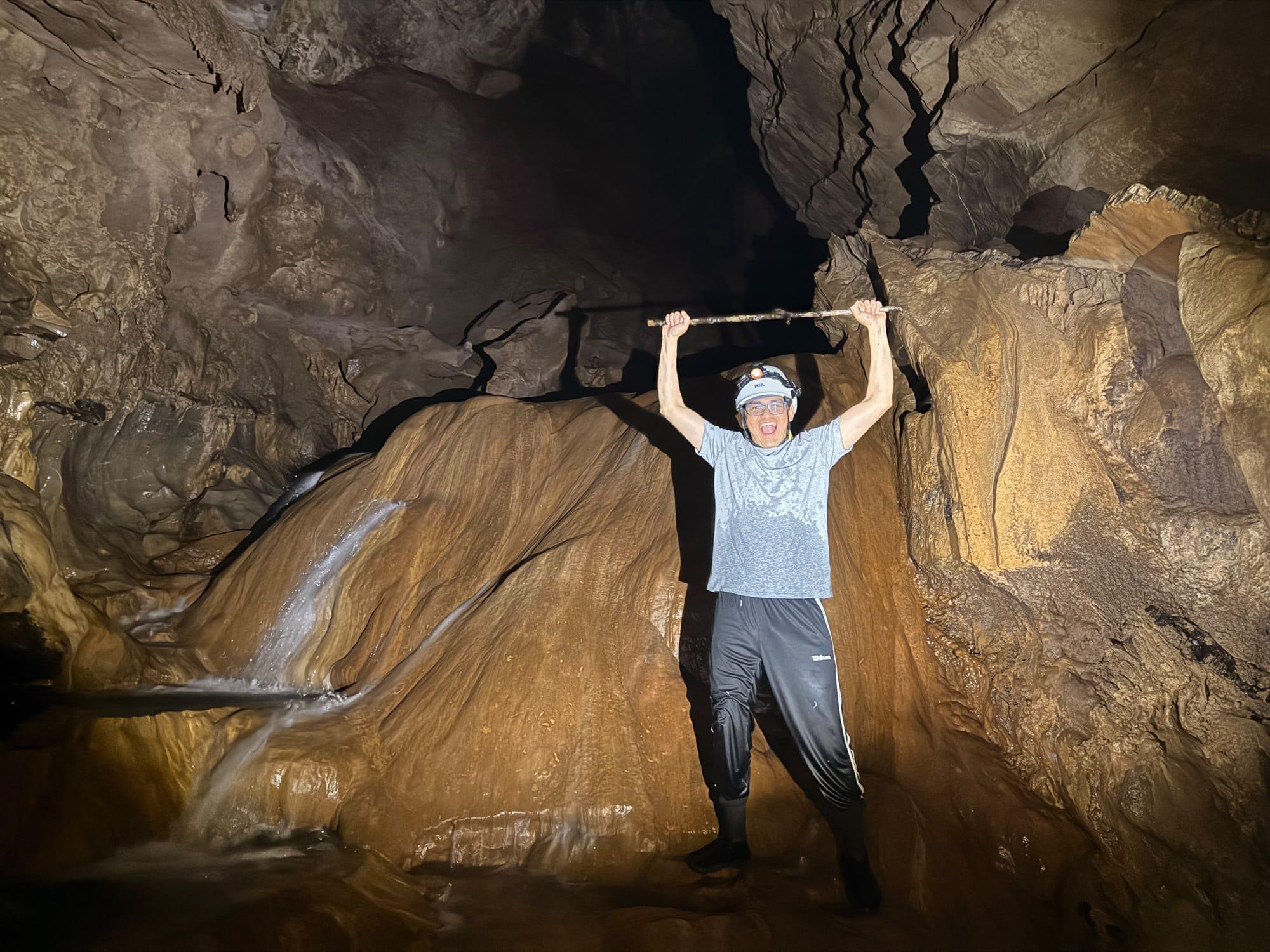 Xilo Cave — arms up in front of a flowstone with water running down it