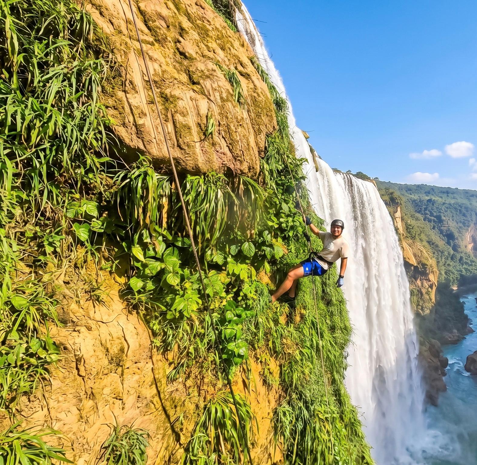 Rapelista apoyado en la pared tapizada de vegetación junto a la cortina de agua de la Cascada de Tamul con el cañón turquesa al fondo
