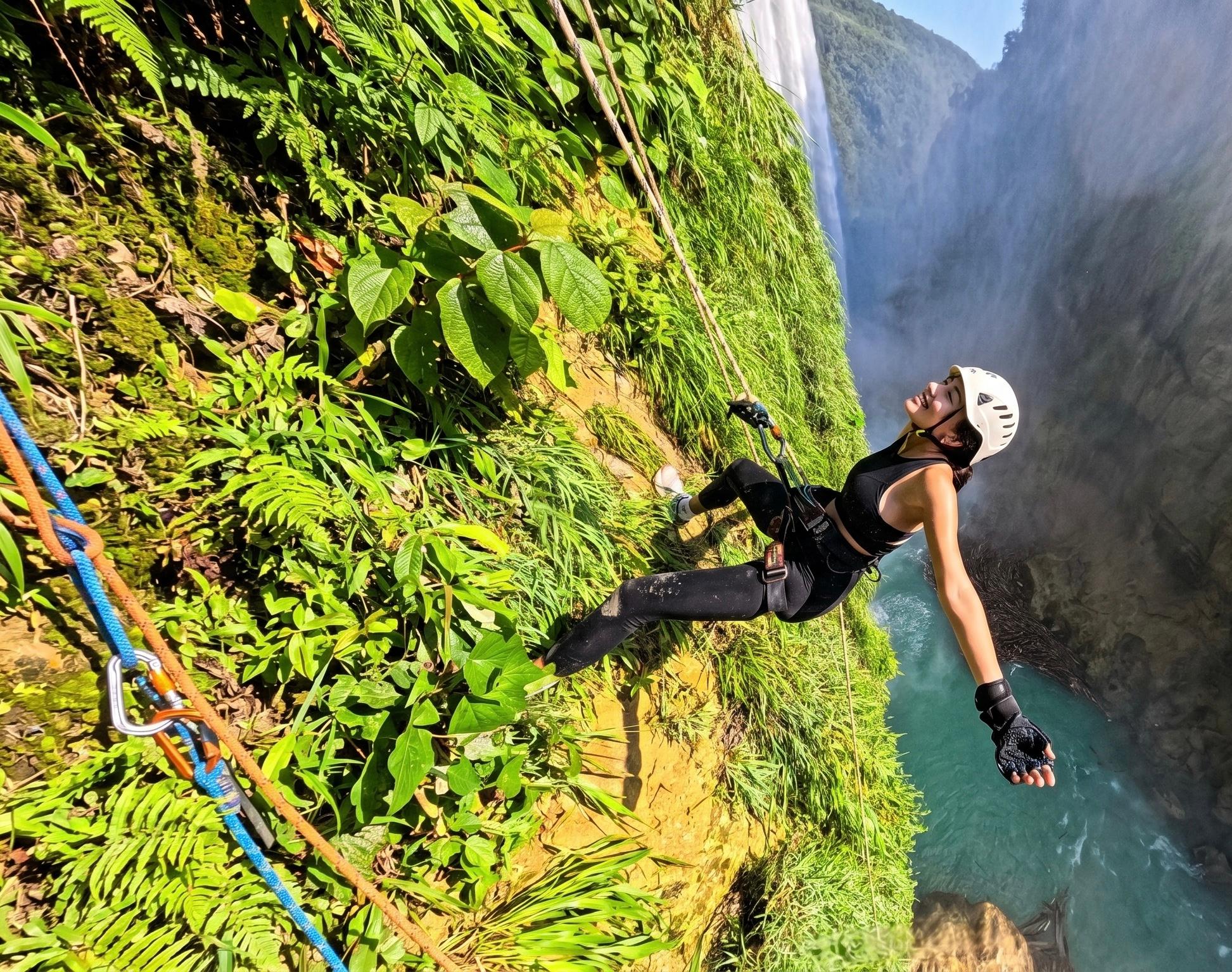 Mujer con casco blanco extendiendo los brazos en rappel sobre el río turquesa del Tampaón con la cascada al fondo