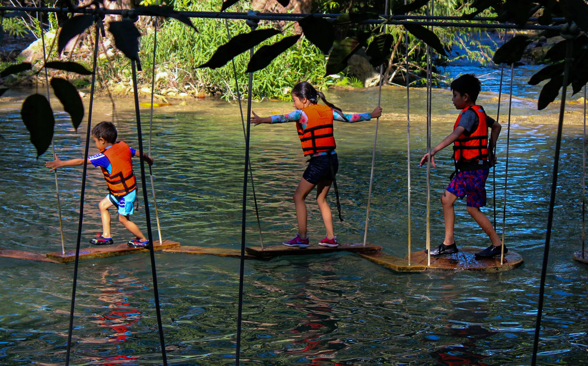 Niños con chalecos salvavidas cruzando un puente colgante sobre río turquesa en Tamasopo — actividades para niños en la Huasteca Potosina